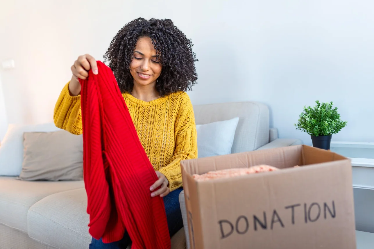 A smiling woman holds up a red knitted sweater beside a box labelled donation